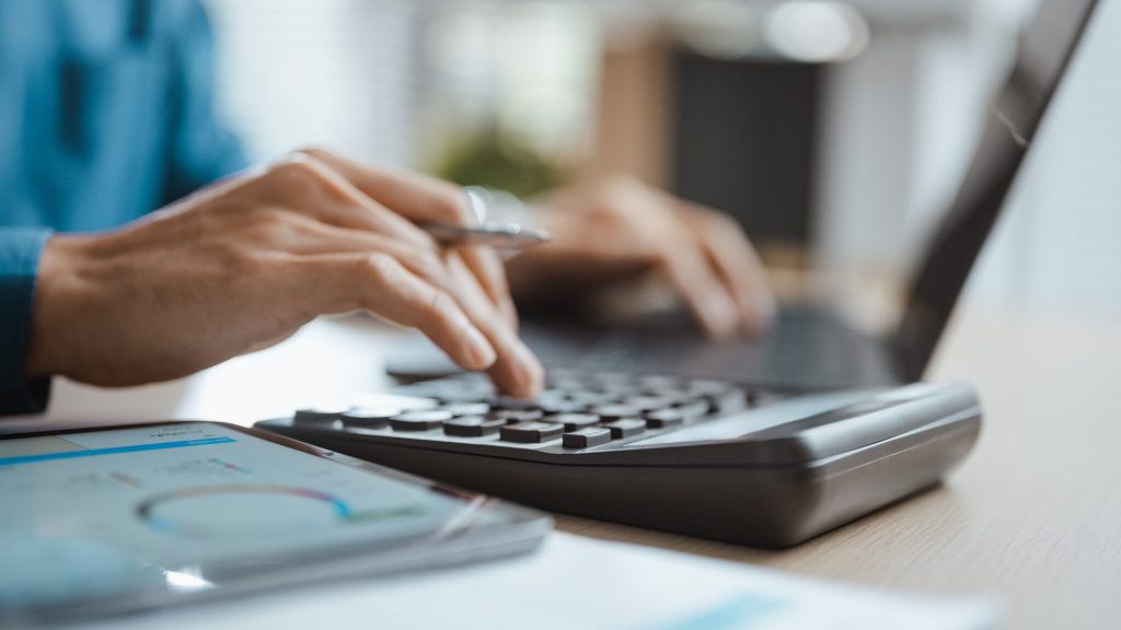 Two colleagues use a calculator and laptop for finance planning, combining budgeting, accounting, expense and cost analysis in a report that helps their business compare revenue and finance goals.