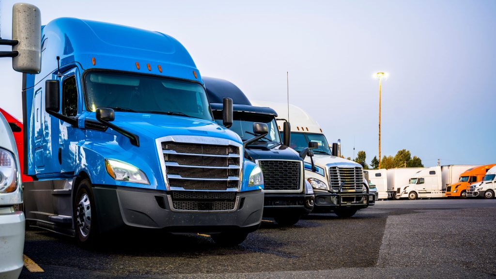 Different makes and models industrial grade professional big rigs semi trucks standing in row on the truck stop parking lot at evening time waiting to continued delivery schedule